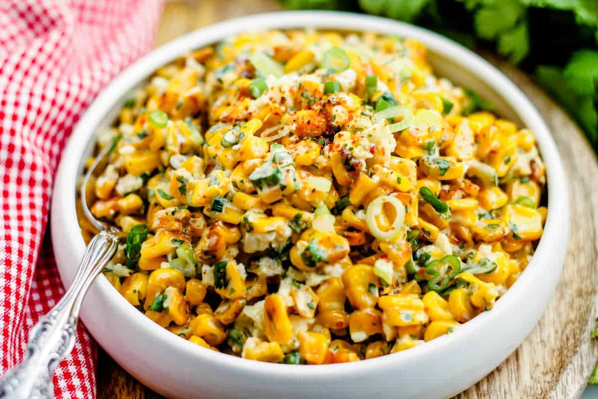 A bowl of Mexican street corn salad topped with chopped green onions, cotija cheese, and spices, served with a spoon.