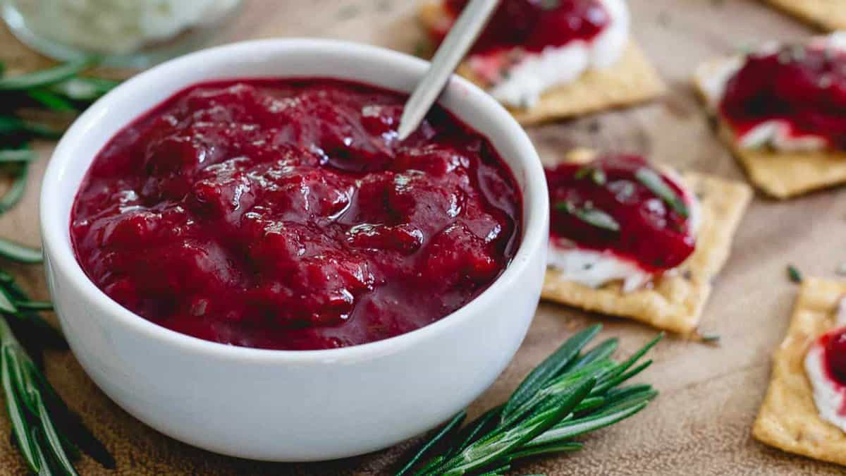 A white bowl filled with cranberry sauce, with a spoon inside. Crackers topped with cranberry sauce and herbs are placed nearby on a wooden surface.