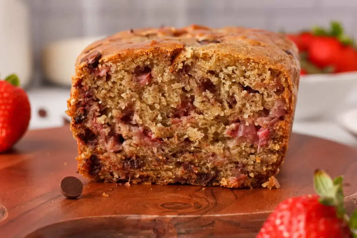 A close-up view of a sliced loaf of strawberry bread on a wooden surface, with fresh strawberries and chocolate chips nearby.
