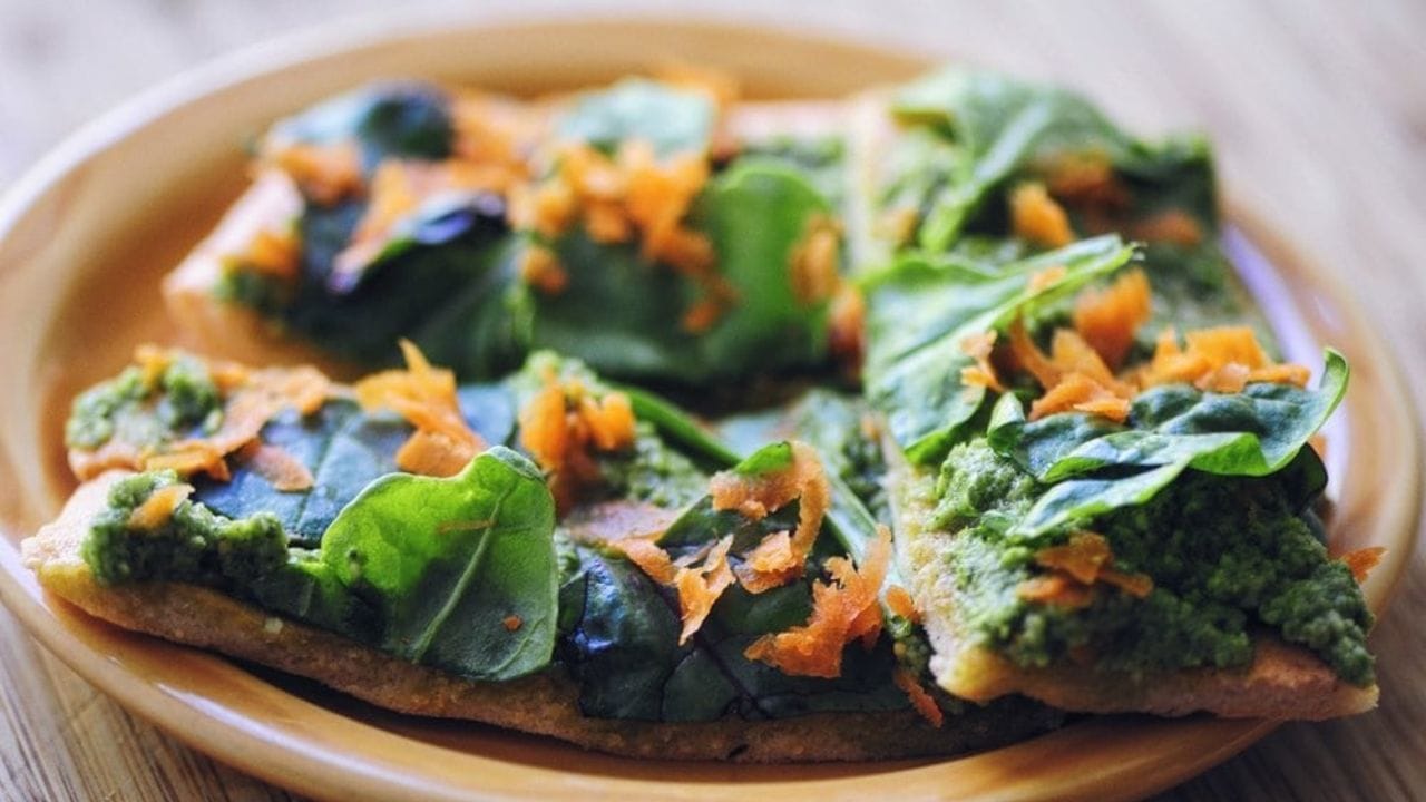 A plate with four slices of flatbread topped with green pesto, fresh spinach leaves, and shredded carrots.