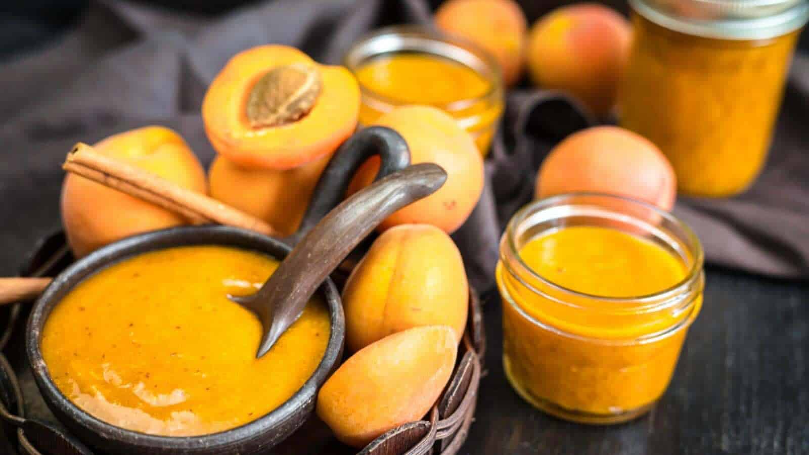 A bowl and jars of apricot sauce with whole and halved apricots, a cinnamon stick, and a spoon, arranged on a dark surface.