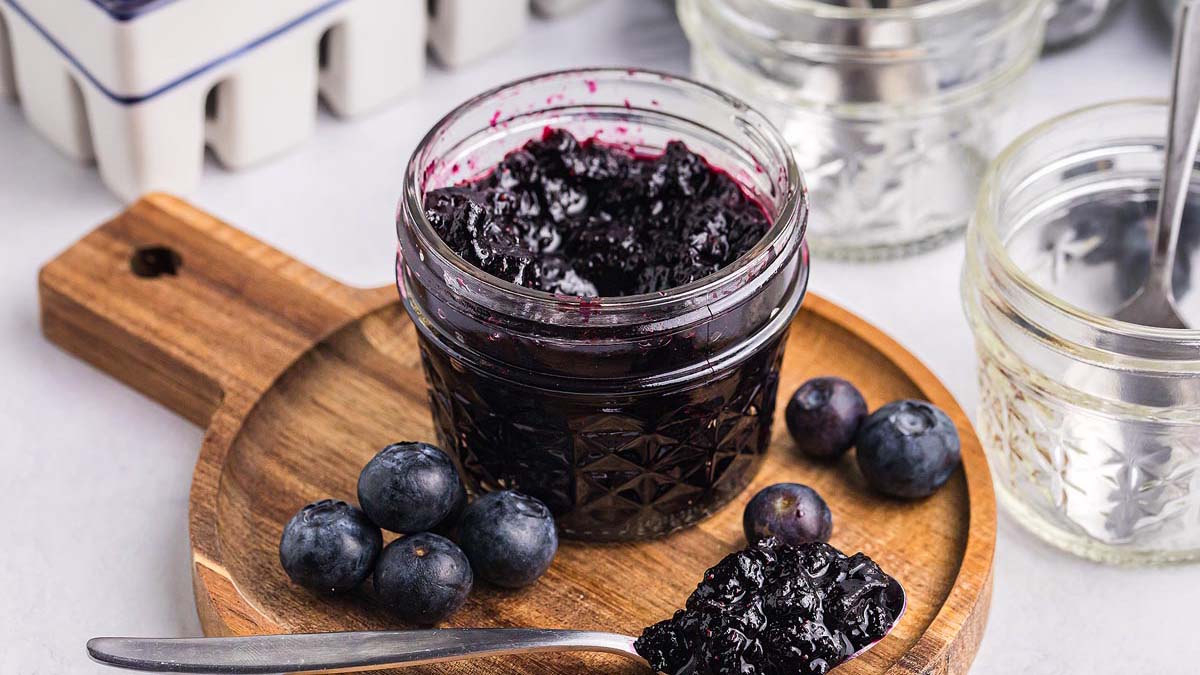 A glass jar of blueberry jam sits on a wooden board with fresh blueberries and a spoonful of jam beside it; empty jars and utensils are in the background.
