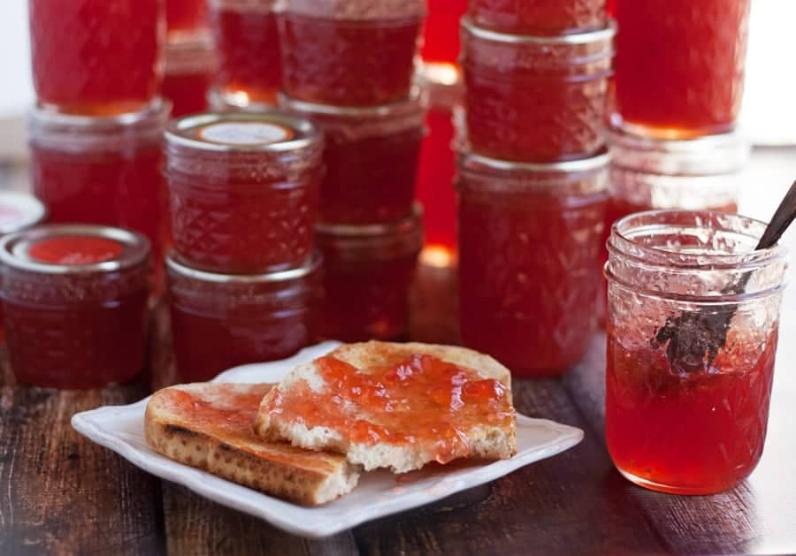 Several jars of red jam are stacked on a wooden table. In the foreground, toast with jam sits on a plate next to an open jar with a spoon inside.