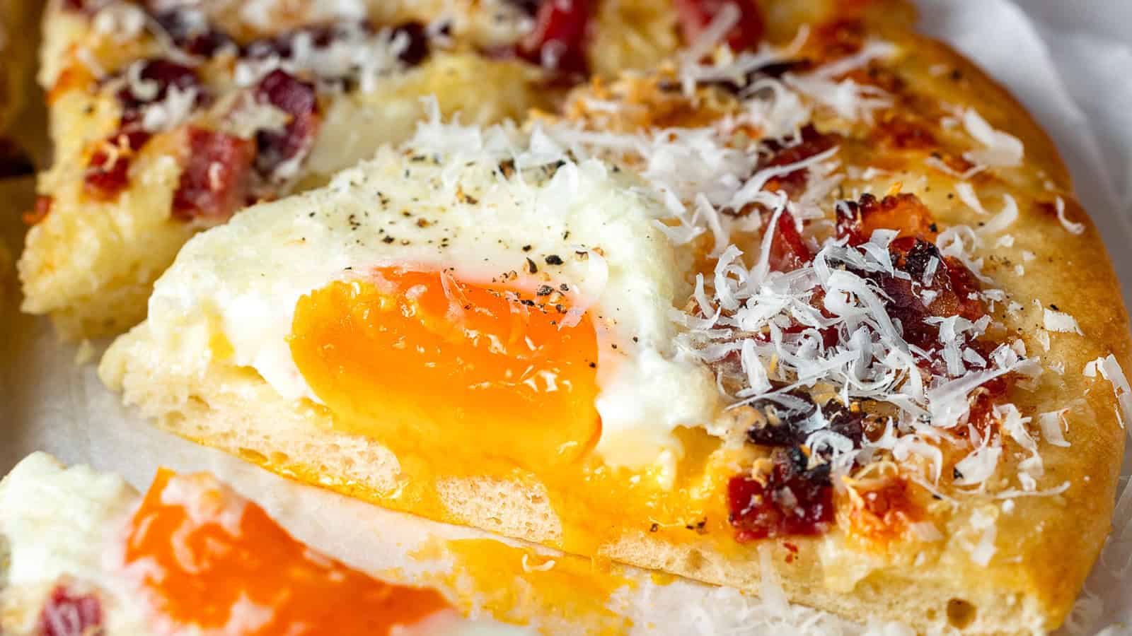 A close-up of a breakfast pizza topped with a runny egg, grated cheese, and pieces of bacon on a golden crust.