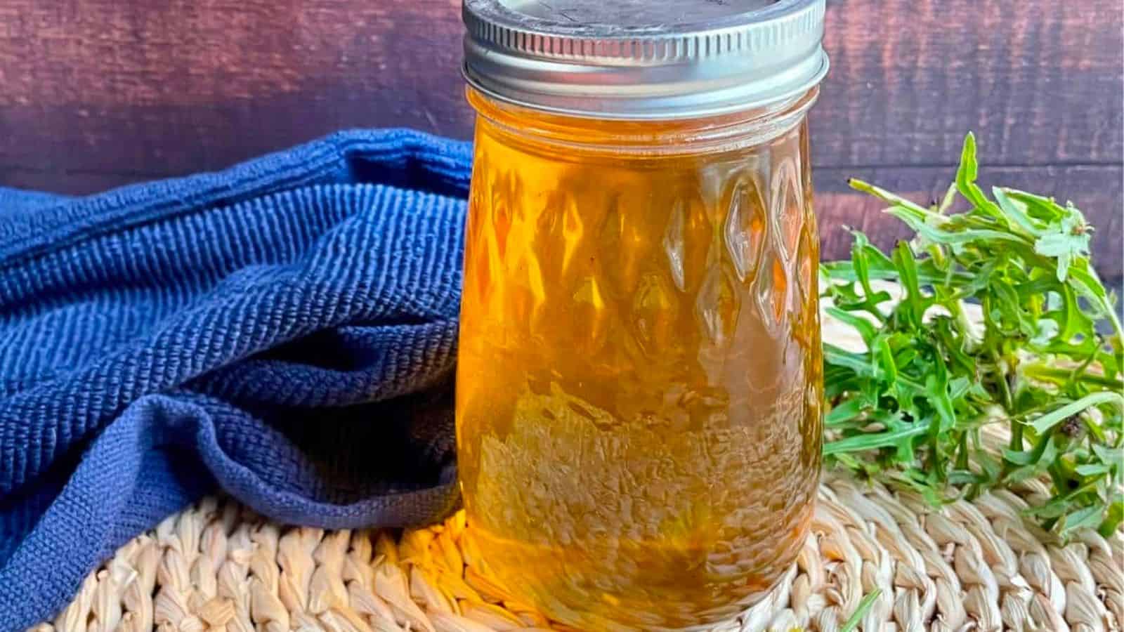 A mason jar filled with golden liquid sits on a woven mat next to a blue cloth and green herbs, against a dark wooden background.