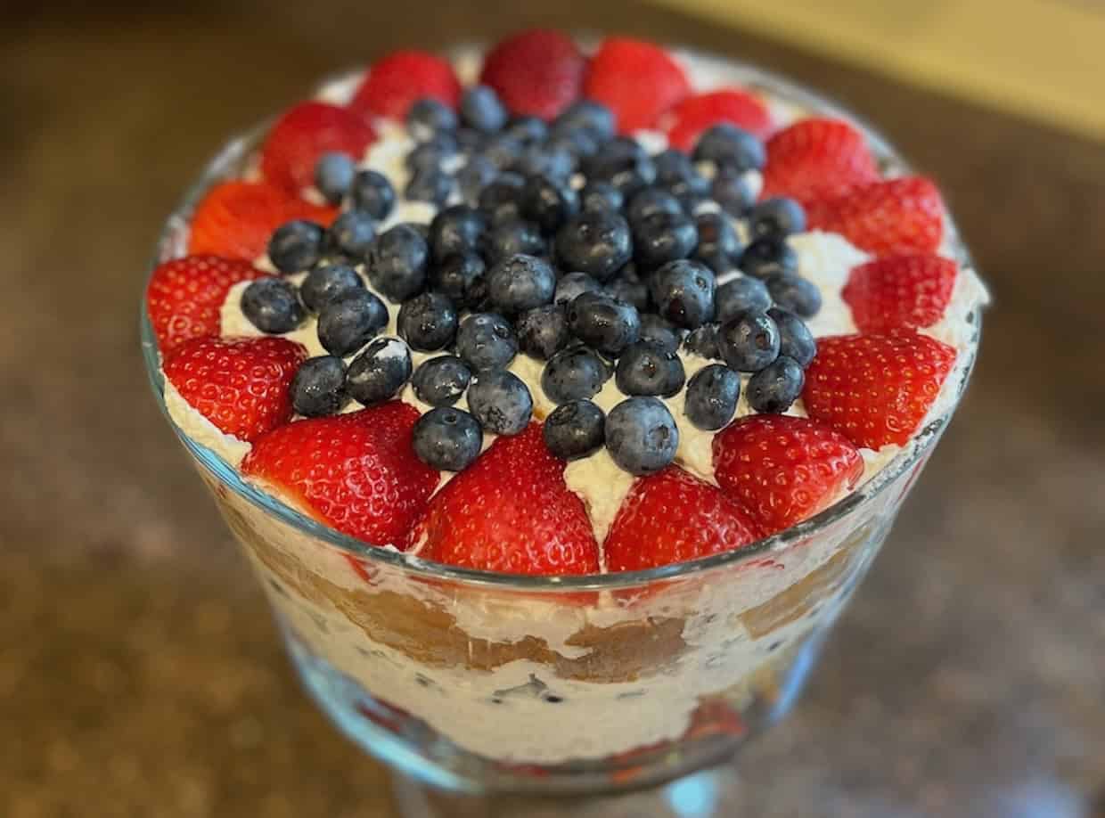 A layered trifle dessert in a glass bowl, topped with whole strawberries around the edge and blueberries in the center.
