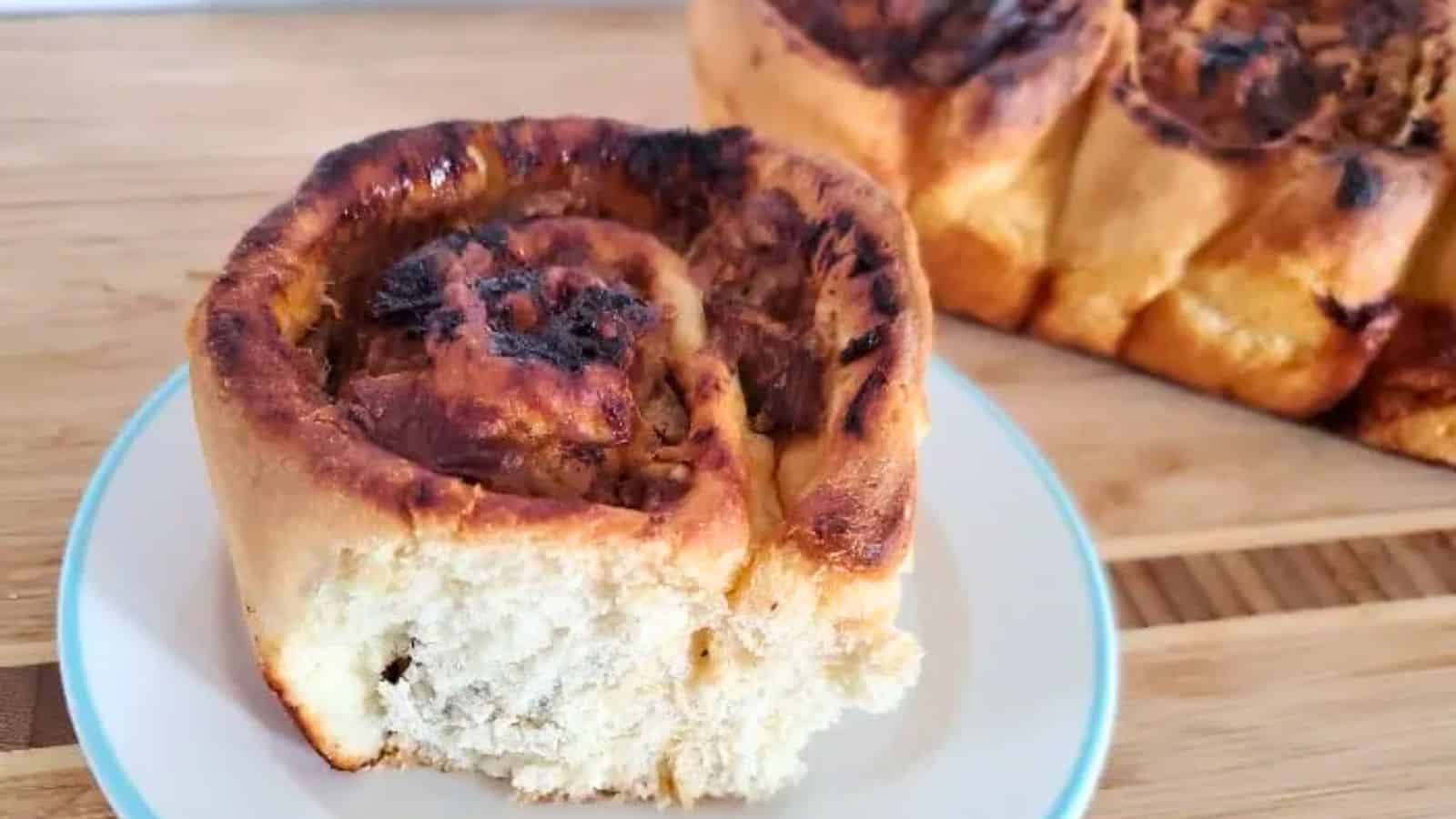 A close-up of a cinnamon roll on a white plate, with more cinnamon rolls in the background on a wooden surface.