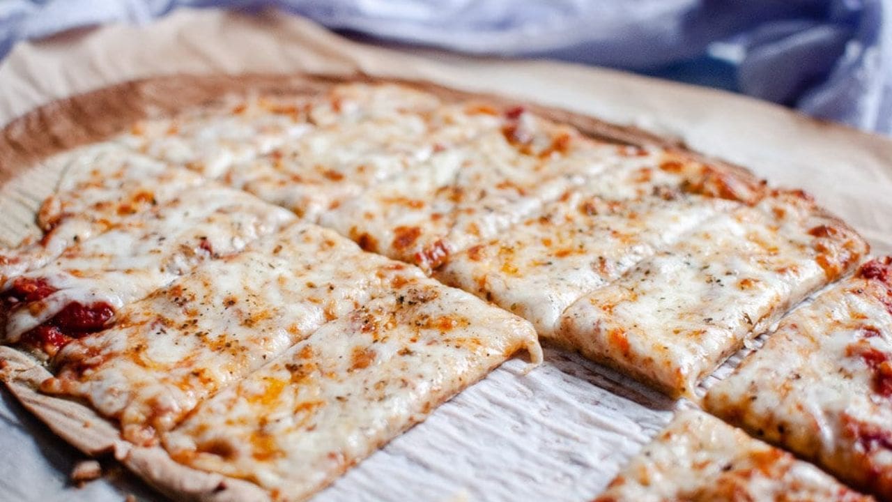 Rectangular thin-crust cheese pizza sliced into squares, topped with melted cheese and herbs, on a sheet of parchment paper.