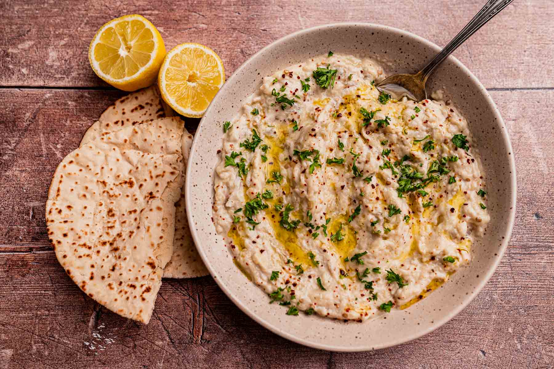 A bowl of baba ganoush garnished with chopped parsley and olive oil, served with pita bread and halved lemons on a rustic wooden surface.