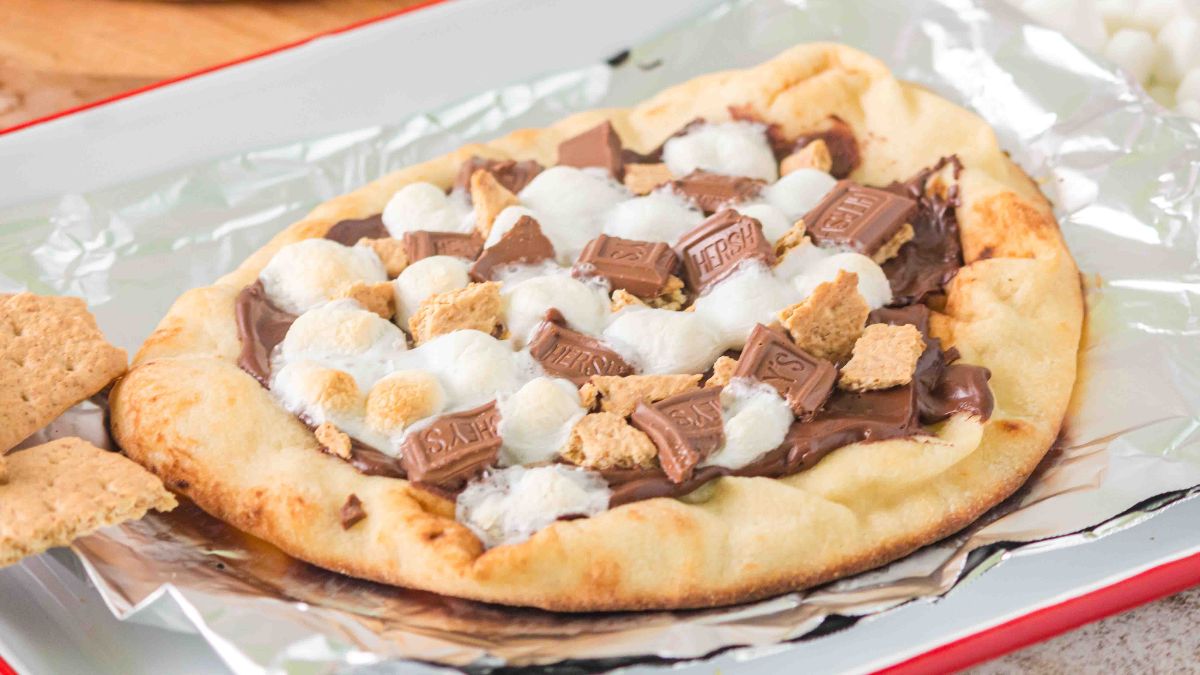A flatbread topped with melted chocolate, mini marshmallows, graham cracker pieces, and chocolate bar chunks, served on foil.
