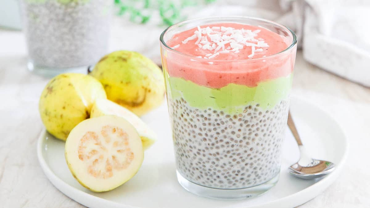 A glass of chia pudding topped with green and pink fruit puree layers and shredded coconut, next to whole and halved guavas on a white plate with a spoon.
