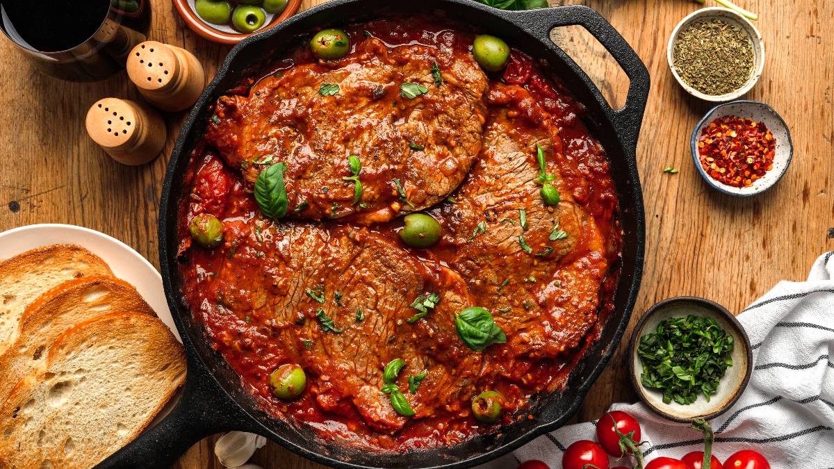 Slices of cooked beef in tomato sauce with green olives and herbs in a cast iron skillet, surrounded by bread, spices, and fresh ingredients on a wooden table.