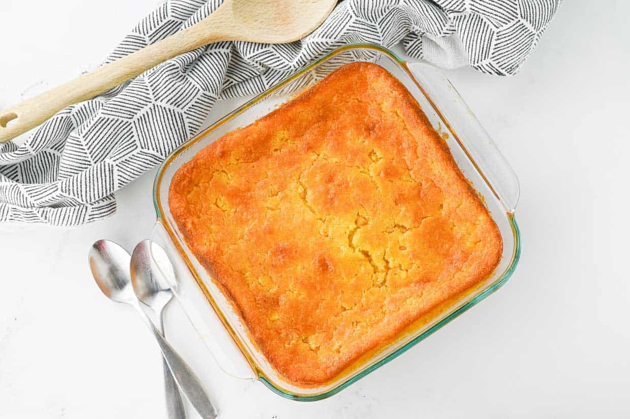A baked cornbread in a square glass dish sits on a white surface next to two metal spoons, wooden utensils, and a patterned cloth.
