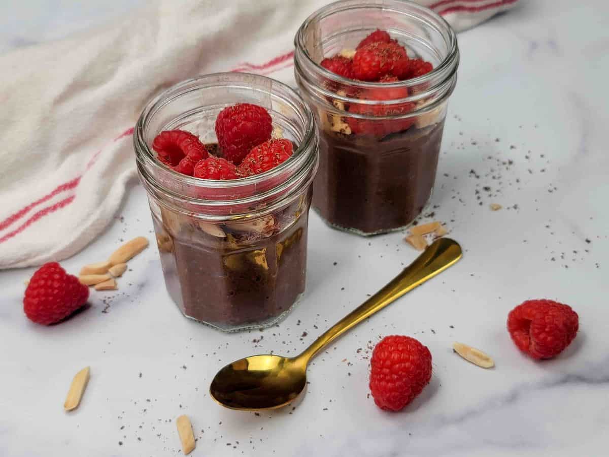 Two glass jars filled with chocolate pudding, topped with raspberries and nuts, sit on a marble surface with a gold spoon and scattered raspberries and nuts nearby.