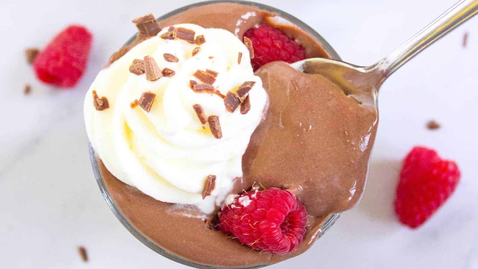 A glass dish of chocolate mousse topped with whipped cream, chocolate shavings, and raspberries, with a spoon scooping some mousse.