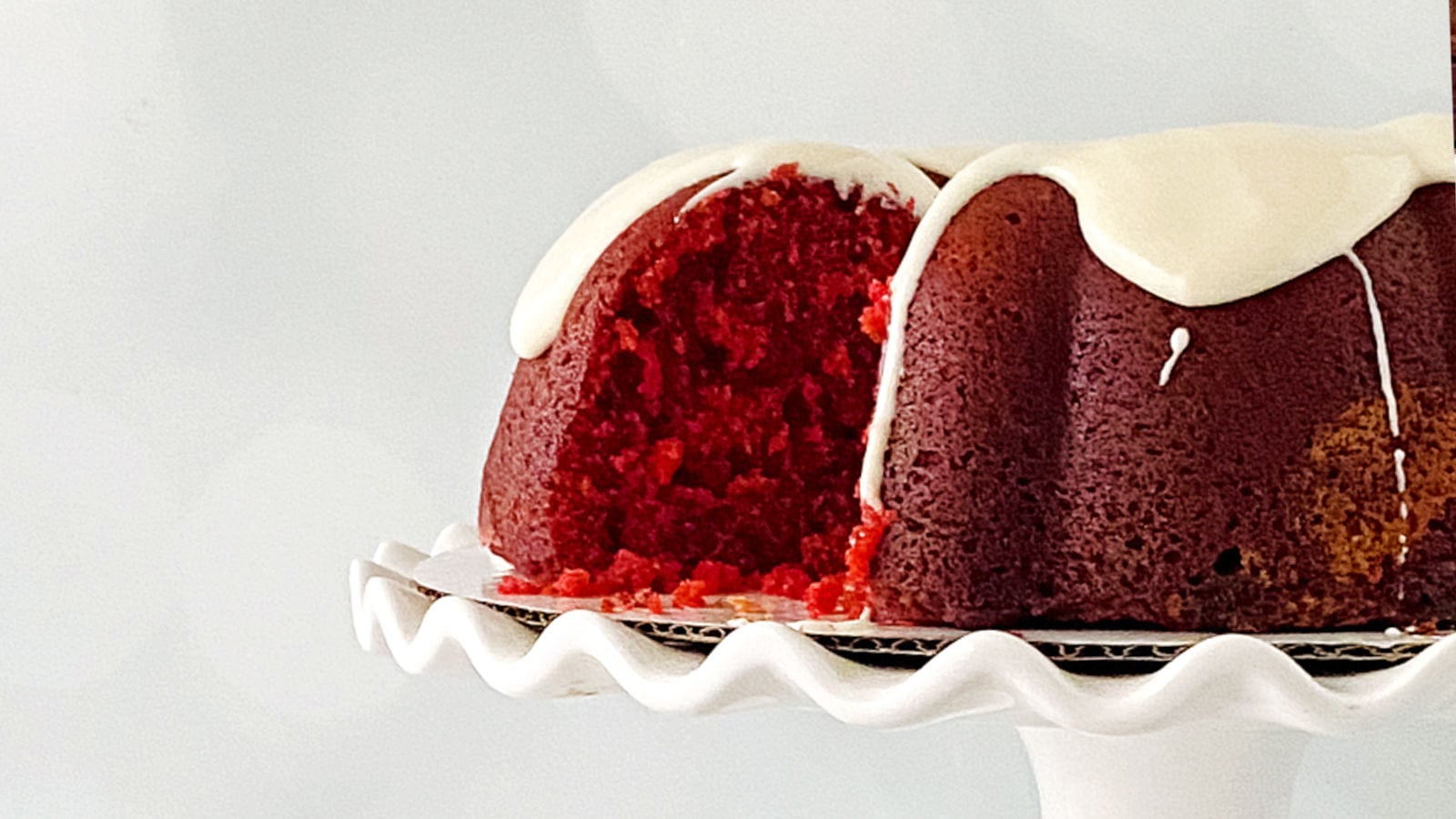 A red velvet bundt cake with white icing, sliced to show the inside, sits on a white cake stand against a plain background.