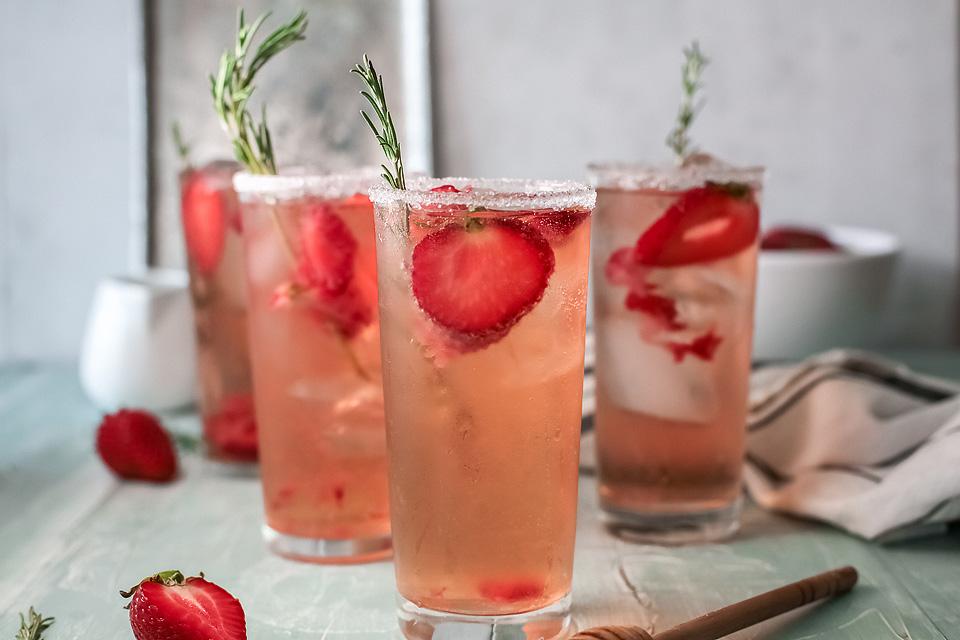 Four glasses of iced strawberry drinks garnished with fresh strawberry slices and rosemary sprigs on a light blue table.