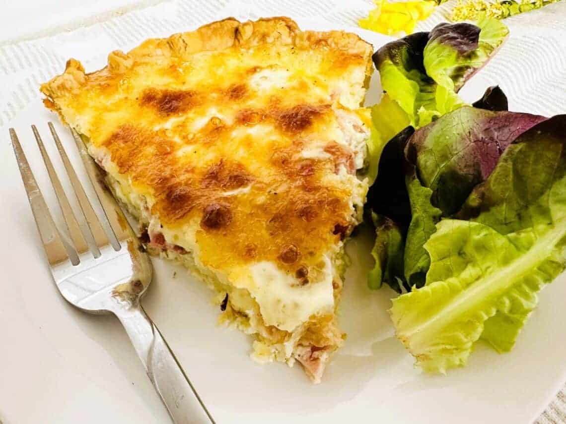 A slice of quiche with a golden-brown top is served on a white plate with mixed leafy greens and a fork.