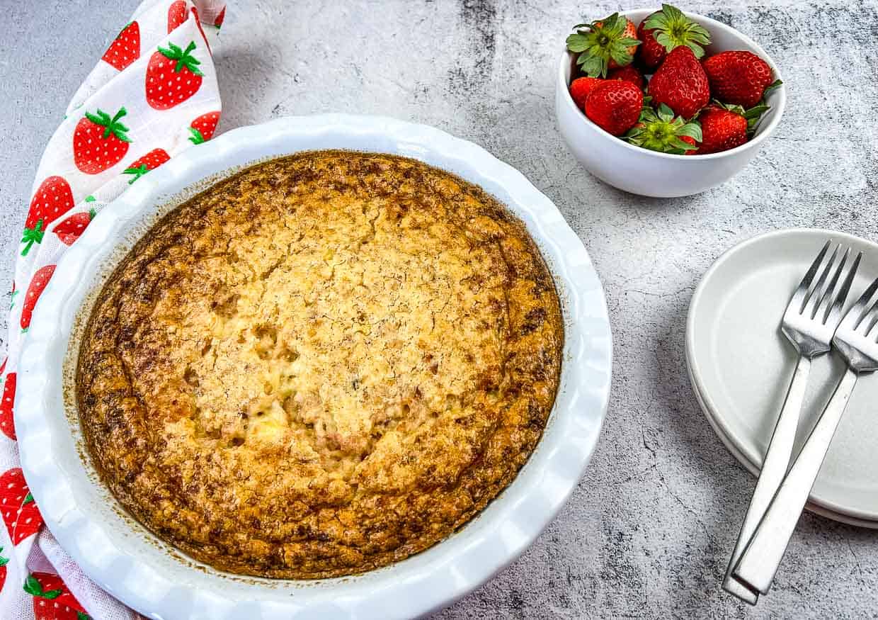 A baked custard pie in a white dish sits next to a bowl of fresh strawberries, a stack of white plates, and three forks on a grey surface.
