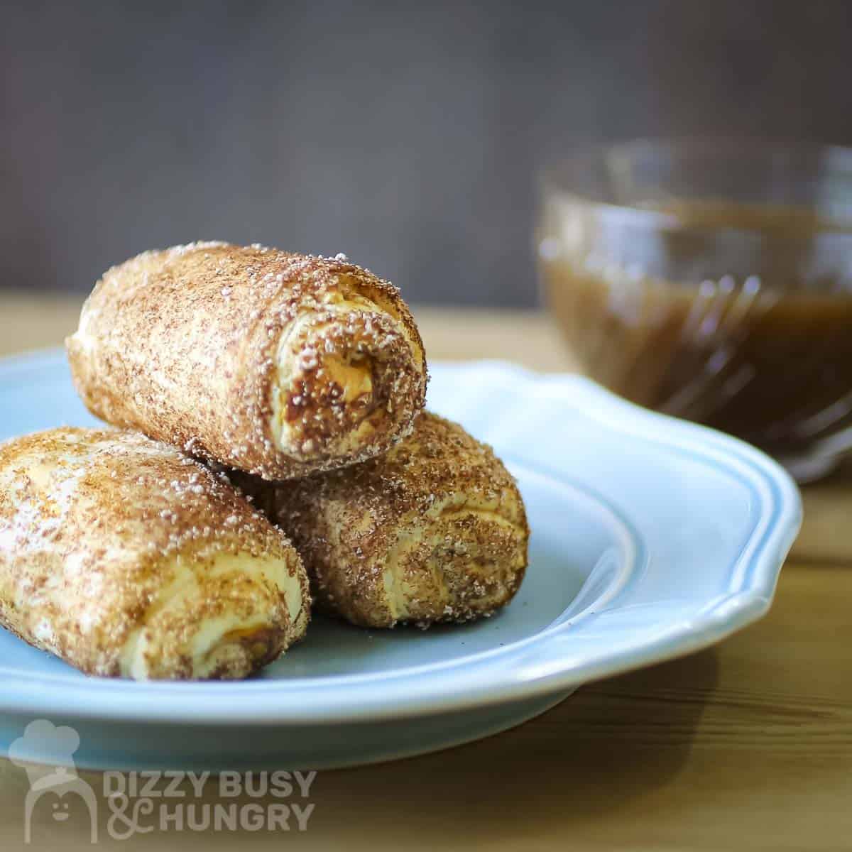 Three cinnamon sugar-coated pastry rolls stacked on a light blue plate, with a glass bowl of brown sauce in the background.