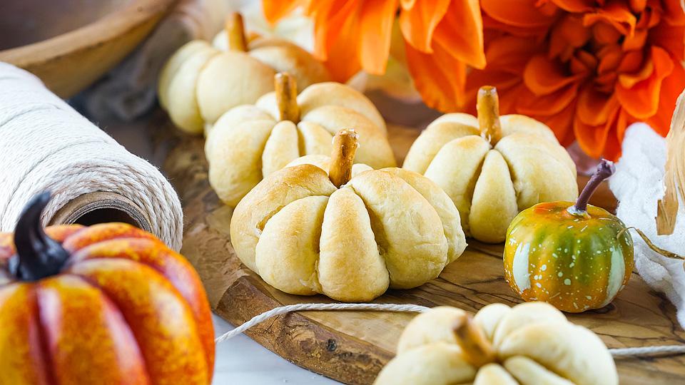 Small pumpkin-shaped bread rolls arranged on a wooden board, surrounded by orange flowers, decorative pumpkins, and a spool of string.