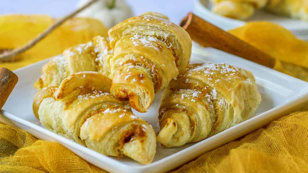 Four golden-brown croissants dusted with powdered sugar are arranged on a white rectangular plate with a yellow cloth underneath.