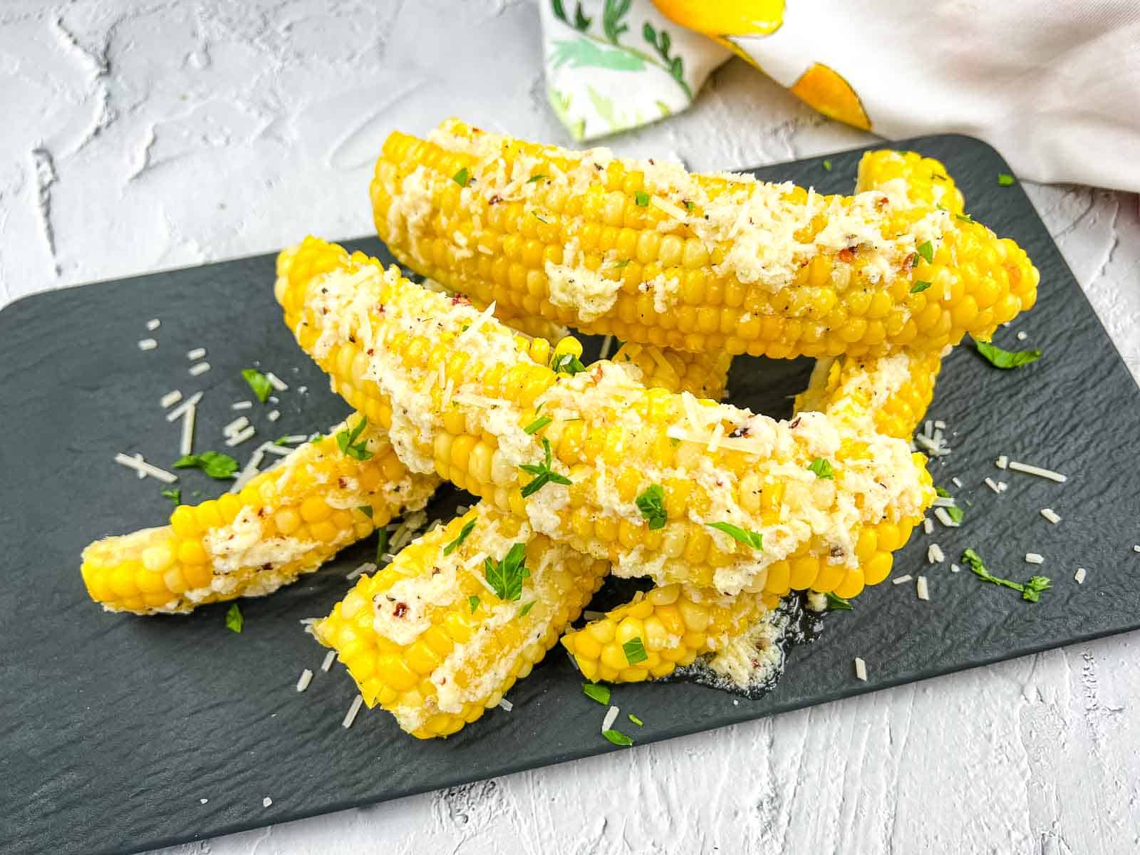 Six pieces of corn on the cob topped with grated cheese and chopped herbs are arranged on a dark slate serving board.