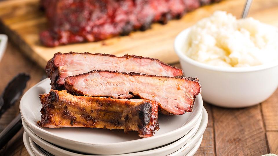 Three slices of barbecue pork ribs are stacked on a plate, with a bowl of mashed potatoes and additional ribs blurred in the background.