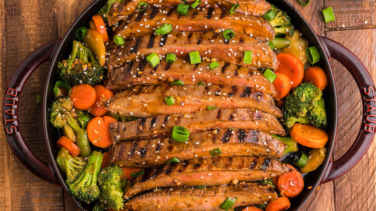 Grilled steak slices topped with sesame seeds and green onions, served over a bed of sautรฉed broccoli and carrots in a round pan on a wooden table.