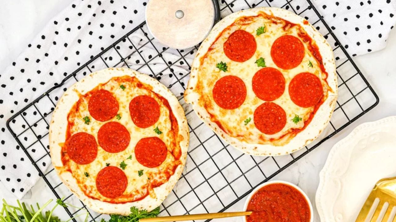 Two small pepperoni pizzas on a cooling rack with a pizza cutter, a bowl of marinara sauce, and a gold fork nearby.