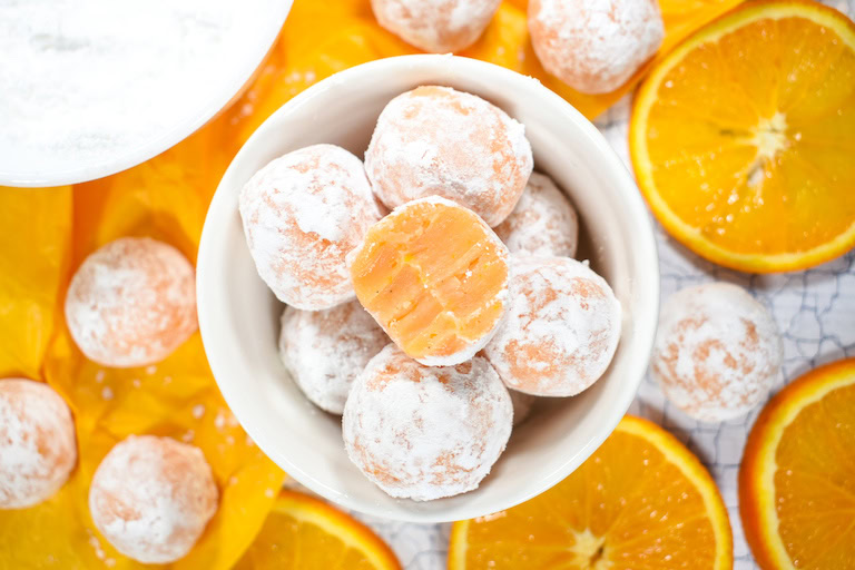 A bowl of powdered sugar-coated orange balls, with one showing an orange filling, surrounded by whole and sliced oranges on a yellow background.