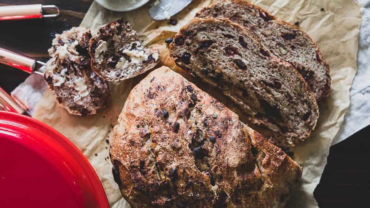 A loaf of rustic bread with visible nuts or fruit is sliced on parchment paper, with one slice spread with butter and a red pot nearby.