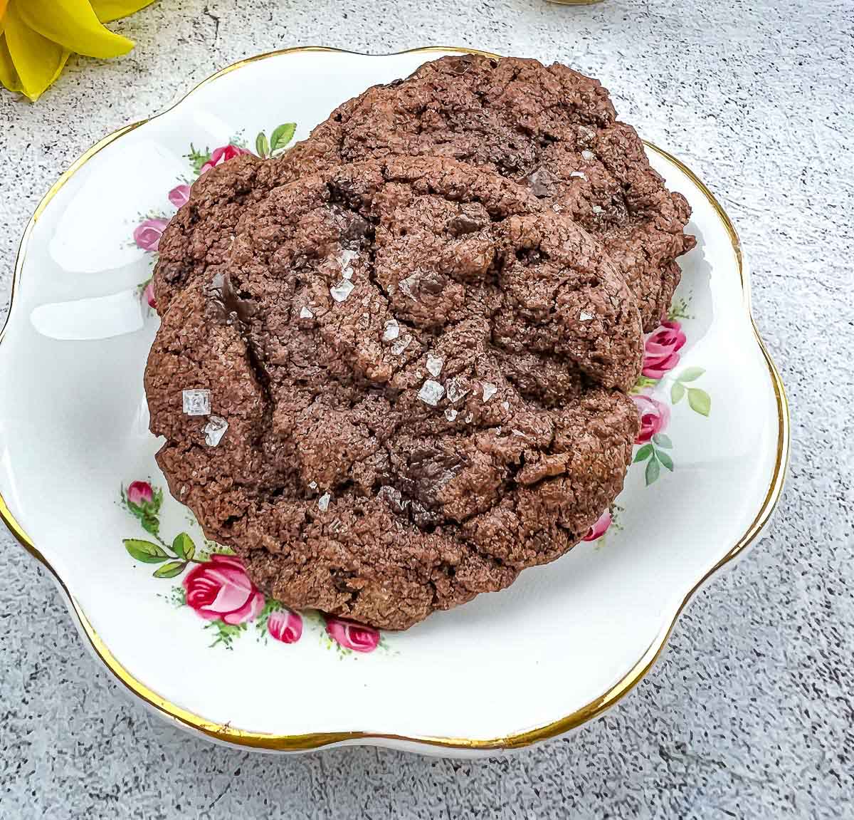 A chocolate cookie topped with sea salt flakes sits on a decorative floral plate against a light textured background.