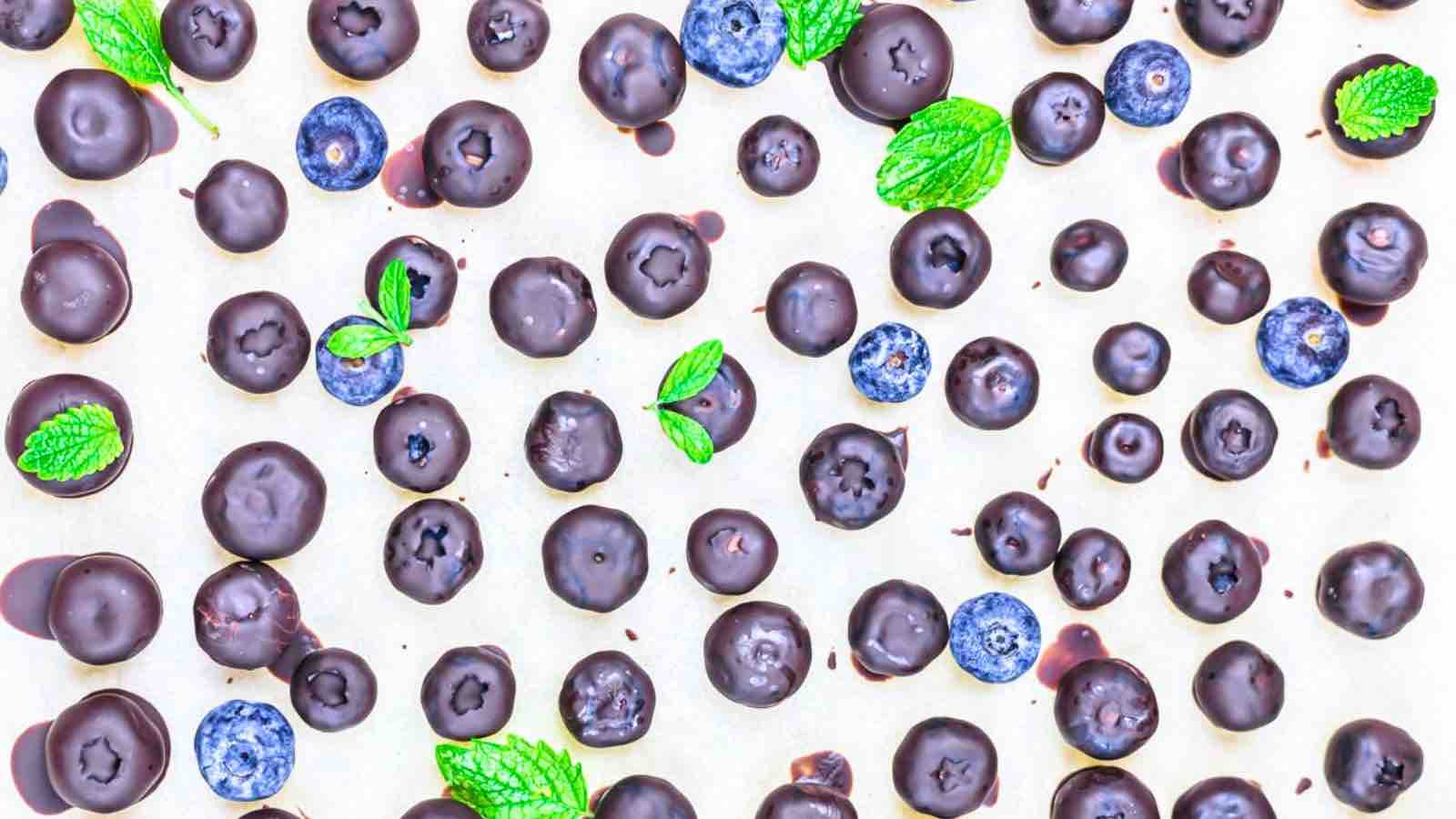 Scattered chocolate-covered blueberries, fresh blueberries, and mint leaves on a light background.