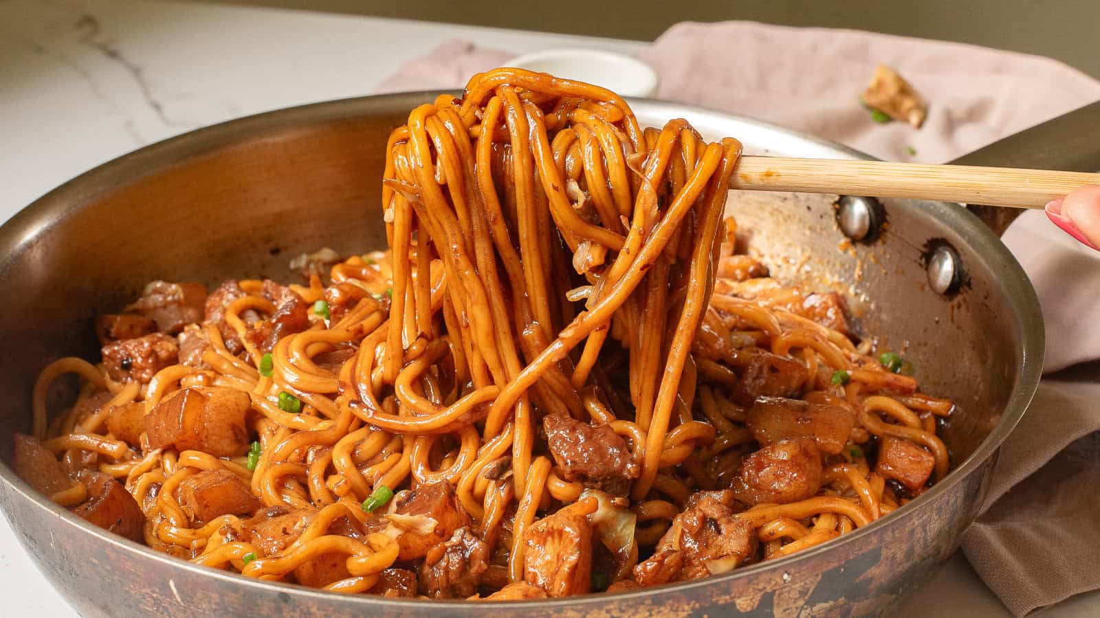 A pan filled with stir-fried noodles, vegetables, and pieces of meat, with a hand lifting some noodles using wooden chopsticks.