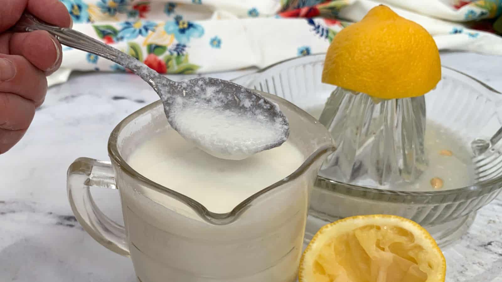A spoon holding a thick white liquid over a glass measuring cup, with a lemon half on a citrus juicer and another squeezed lemon half nearby.