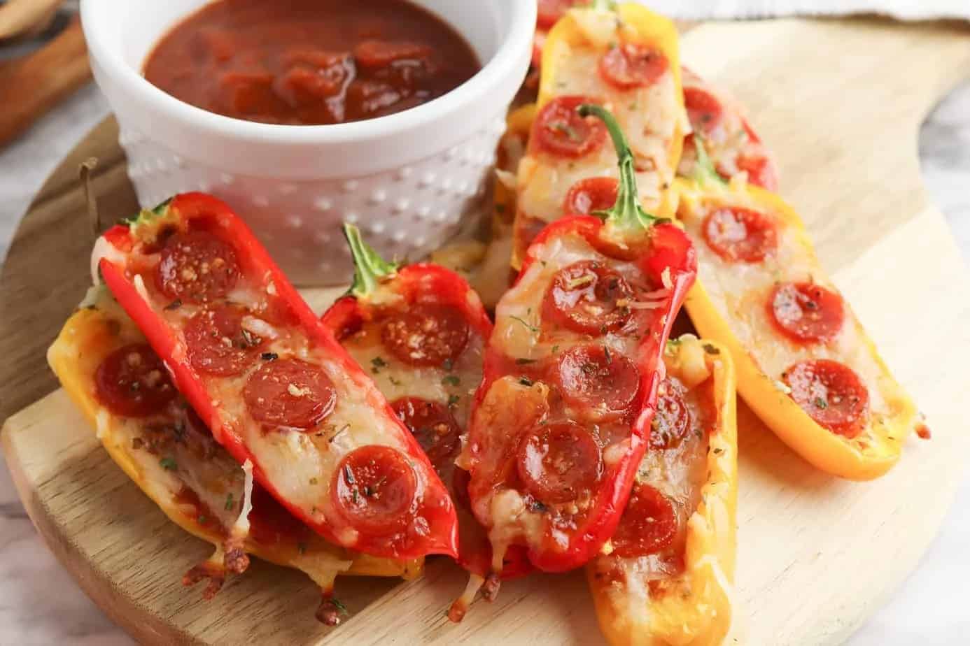 Mini bell peppers stuffed with melted cheese and mini pepperoni slices, served on a wooden board with a small bowl of marinara sauce.