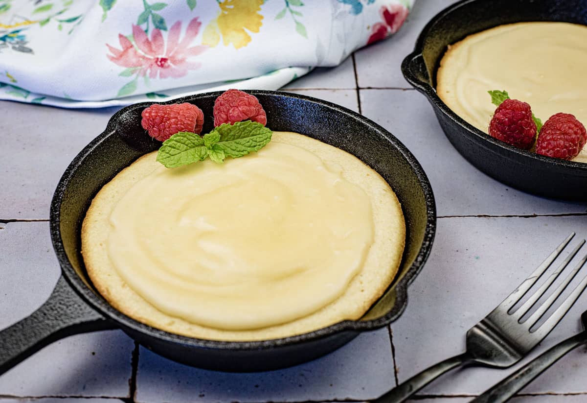 Mini skillet cheesecakes topped with raspberries and mint leaves, placed on a tiled surface with a fork and floral napkin nearby.