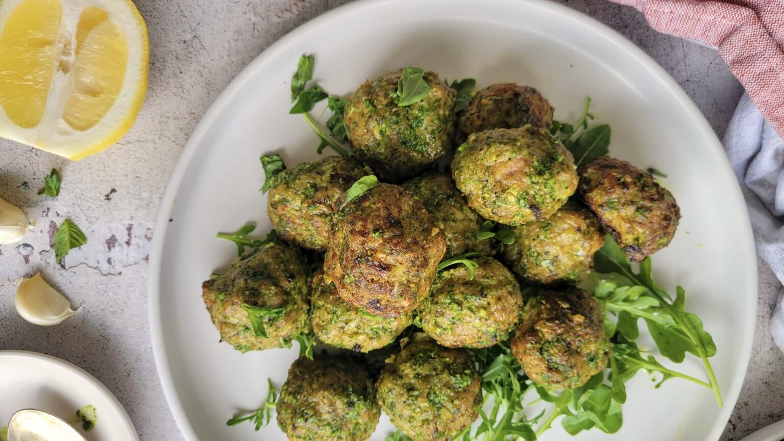 A plate of cooked green herb meatballs garnished with fresh arugula, with a lemon wedge and garlic cloves nearby.
