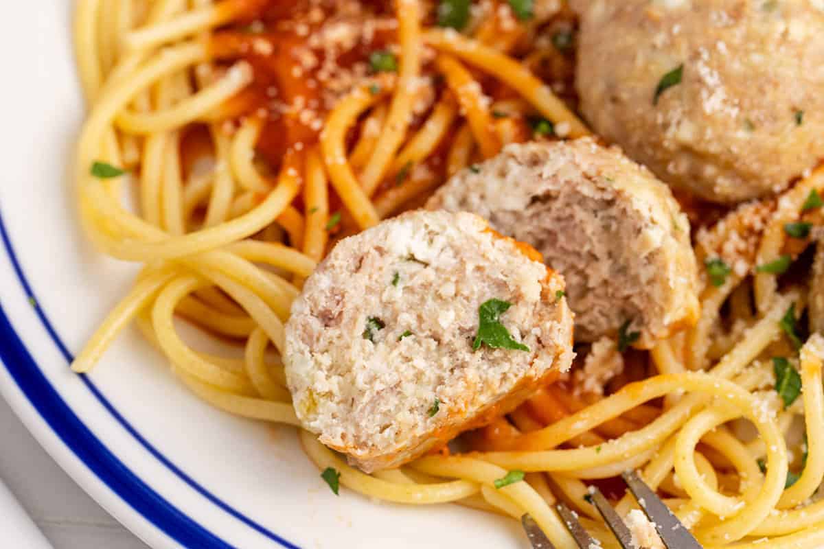 A close-up of spaghetti with tomato sauce and a sliced meatball, garnished with parsley and grated cheese on a white plate with a blue rim.