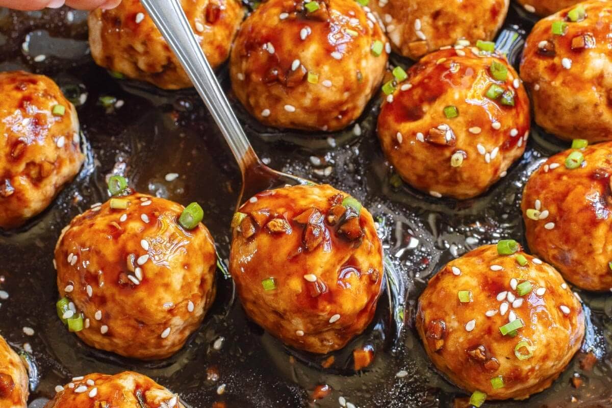 Close-up of glazed meatballs garnished with sesame seeds and chopped green onions, with a spoon lifting one meatball from the sauce.