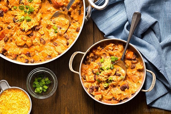 A bowl and pot of cheesy southwest chicken casserole with beans and vegetables; chopped green onions and shredded cheese are in small bowls nearby on a wooden table.