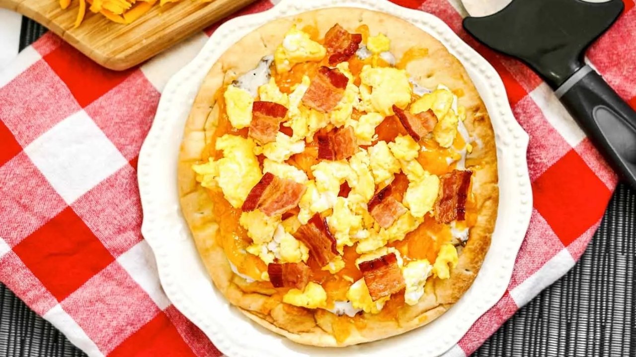 A breakfast pizza topped with scrambled eggs, bacon, and melted cheese sits on a white plate over a red and white checkered tablecloth.