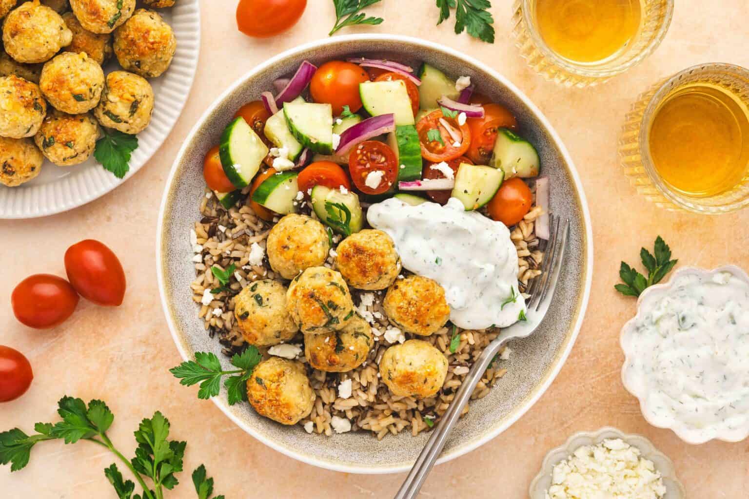 A bowl of rice topped with chicken meatballs, tzatziki sauce, feta cheese, and a Greek salad with cucumbers, tomatoes, and red onions, with drinks and garnishes on the side.