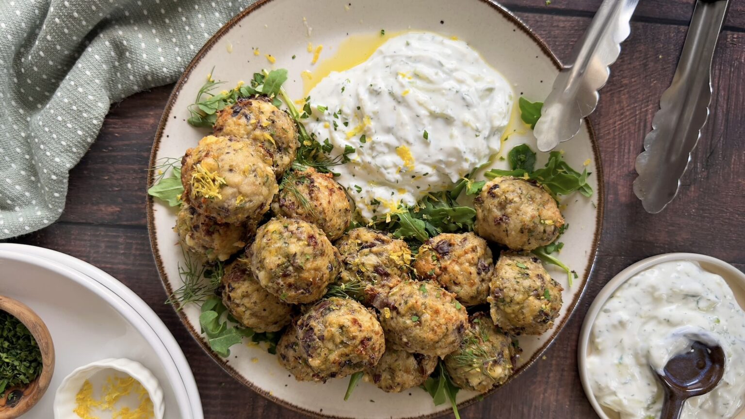 A plate of meatballs served on greens with a side of creamy white sauce, garnished with herbs and lemon zest. Metal tongs and small dishes are visible beside the plate.