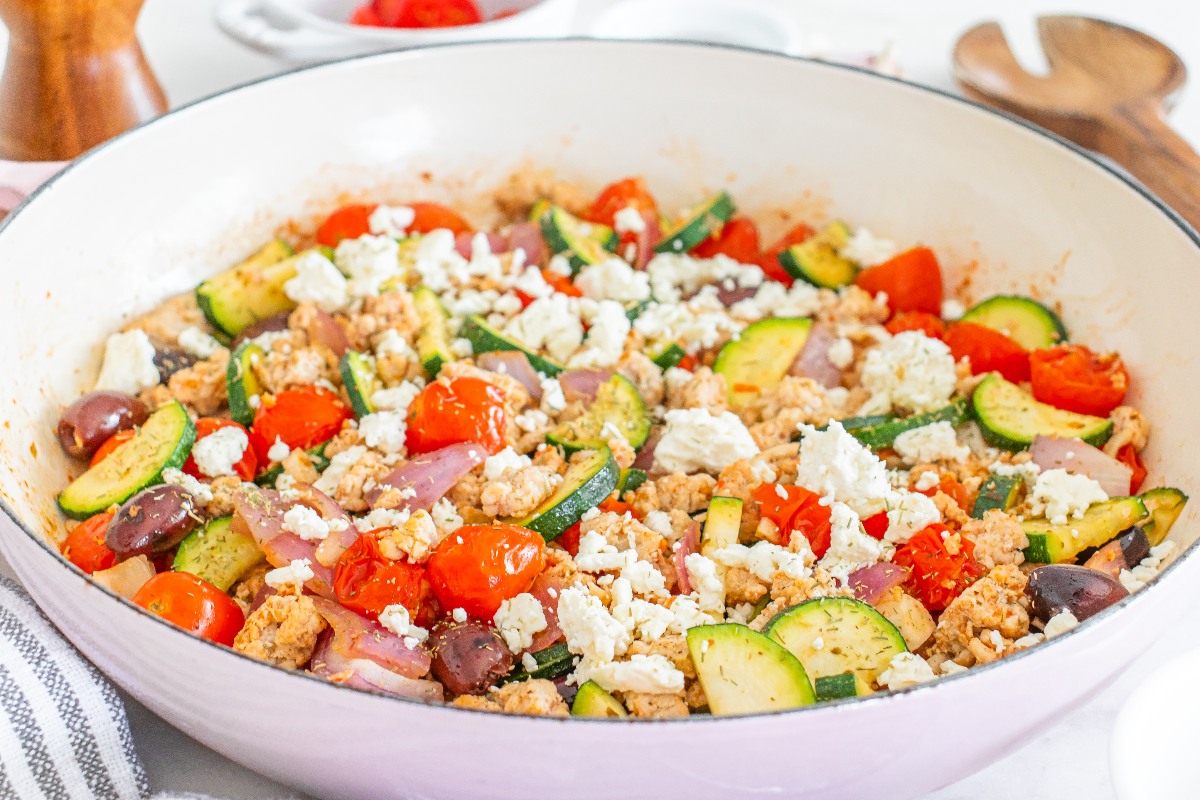 A skillet filled with a mixture of cooked ground meat, zucchini, cherry tomatoes, red onions, olives, and crumbled feta cheese.