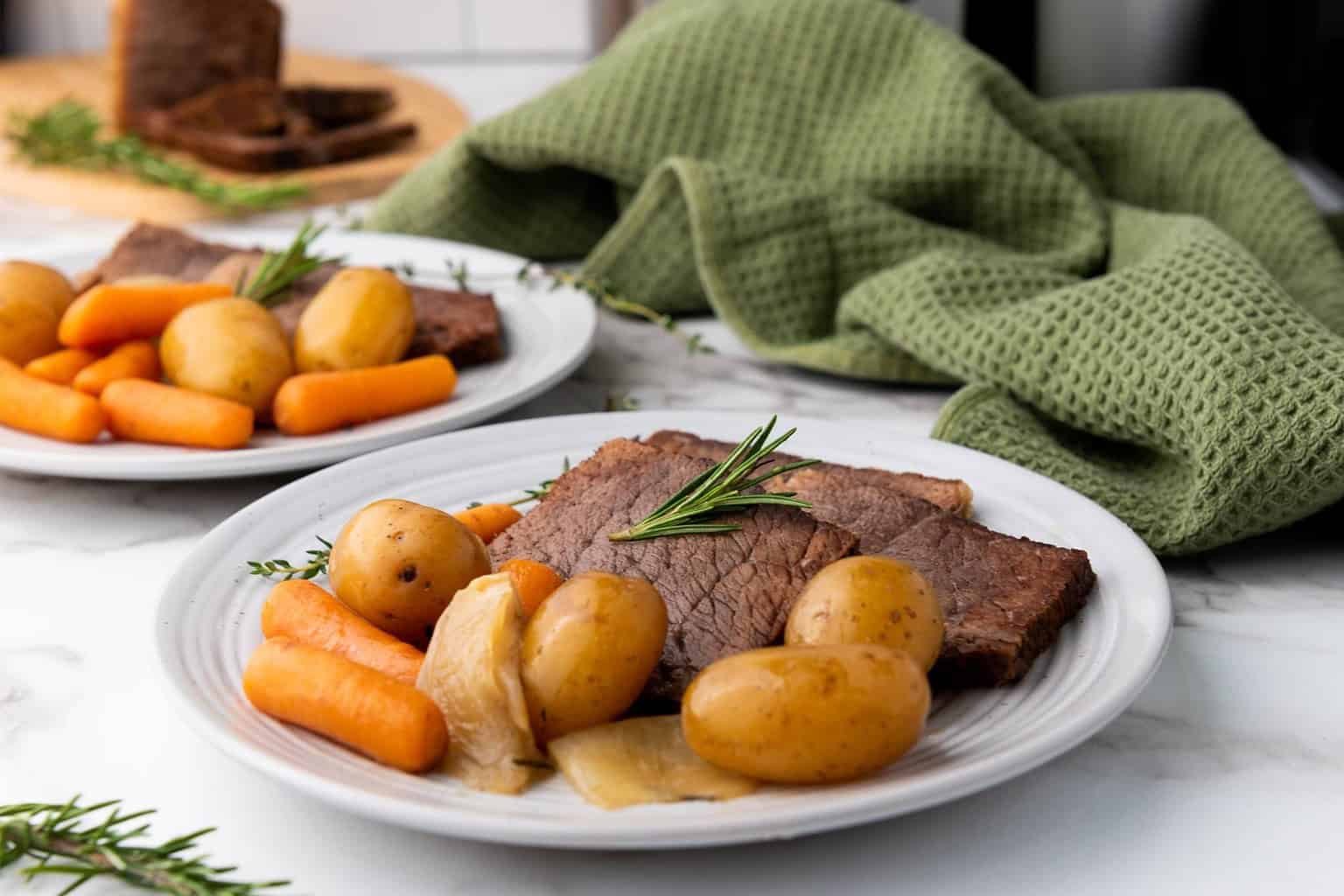 A plate with slices of roast beef, baby potatoes, carrots, and onions, garnished with rosemary, sits on a marble surface next to a green textured cloth.