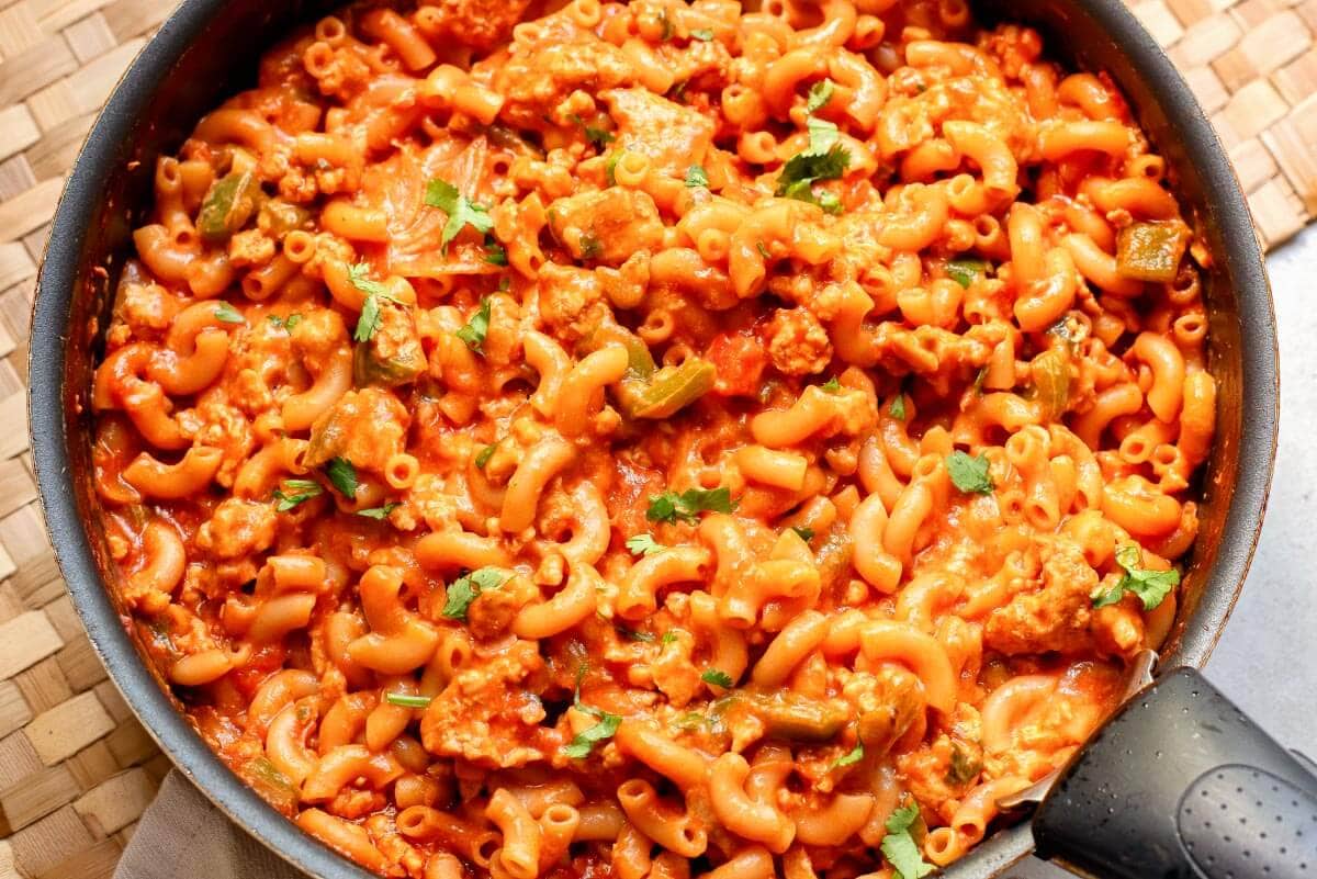 A skillet filled with cooked elbow macaroni mixed with ground meat, tomato sauce, and garnished with chopped herbs.