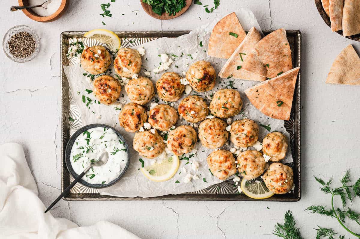 A tray of baked meatballs garnished with herbs and feta, served with pita bread, lemon slices, and a bowl of white dipping sauce.