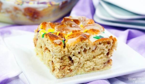 A square slice of iced cinnamon roll cake with sprinkles on a white plate, set on a purple cloth with plates and more cake in the background.